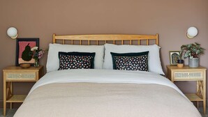 Una cama bien hecha con cabecera de madera, dos mesitas de noche con lámparas y un cuadro enmarcado sobre una de ellas.