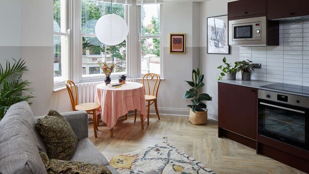 Una cocina moderna con microondas, horno y armarios oscuros. Hay un comedor con una mesa, sillas y un florero con flores. La habitación tiene ventanas grandes con cortinas blancas y vistas a la vegetación.