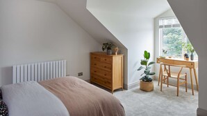 Un dormitorio con techo inclinado, un radiador, un aparador de madera, un escritorio con silla y una ventana con vistas al verdor.