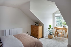Relax in this serene bedroom with a large bed, wooden dresser, and desk near a window offering a view of lush greenery.
