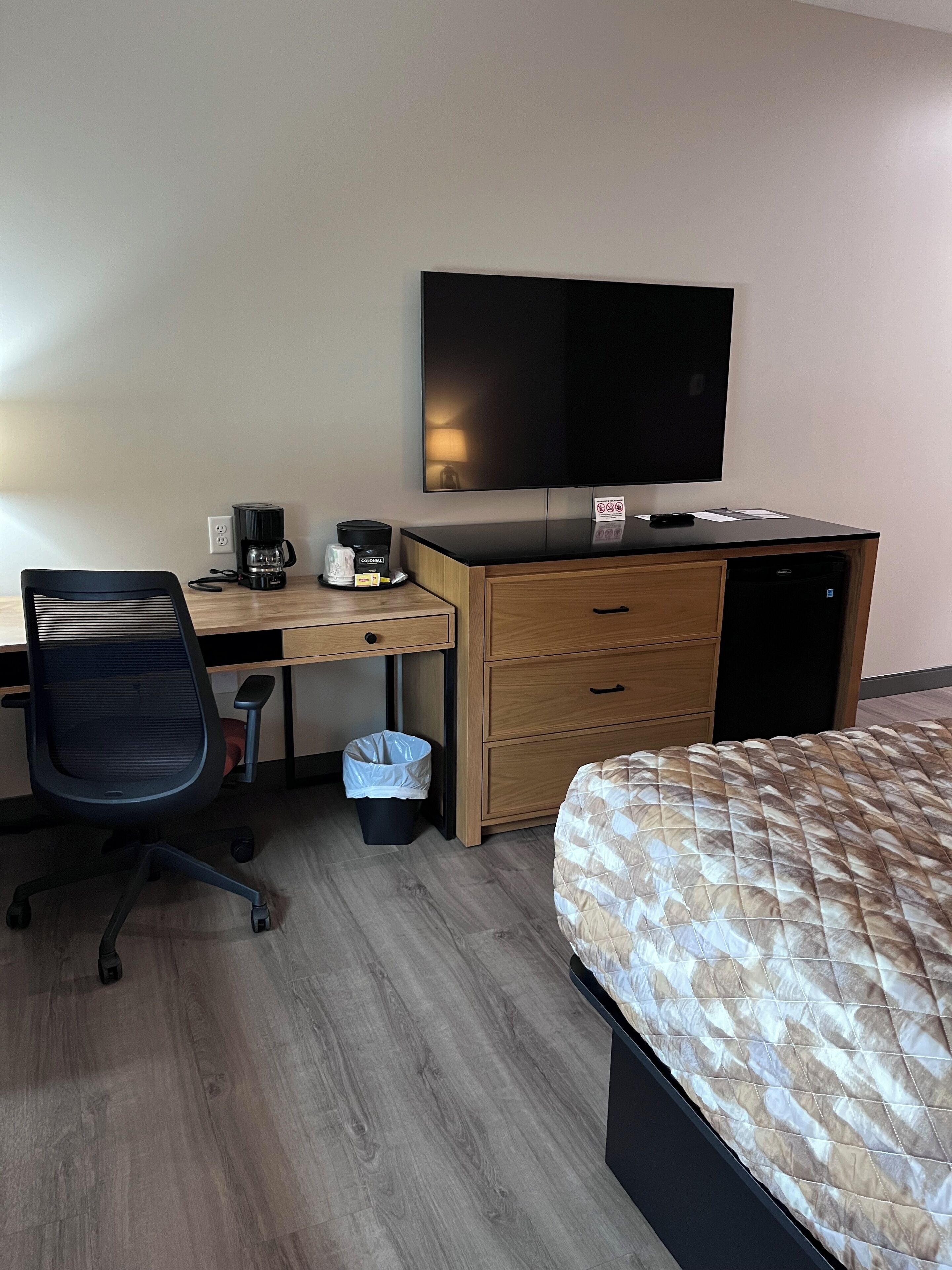 A hotel room with a bed, a desk with a computer, a chair, a coffee maker, a trash bin, and a television on the wall.