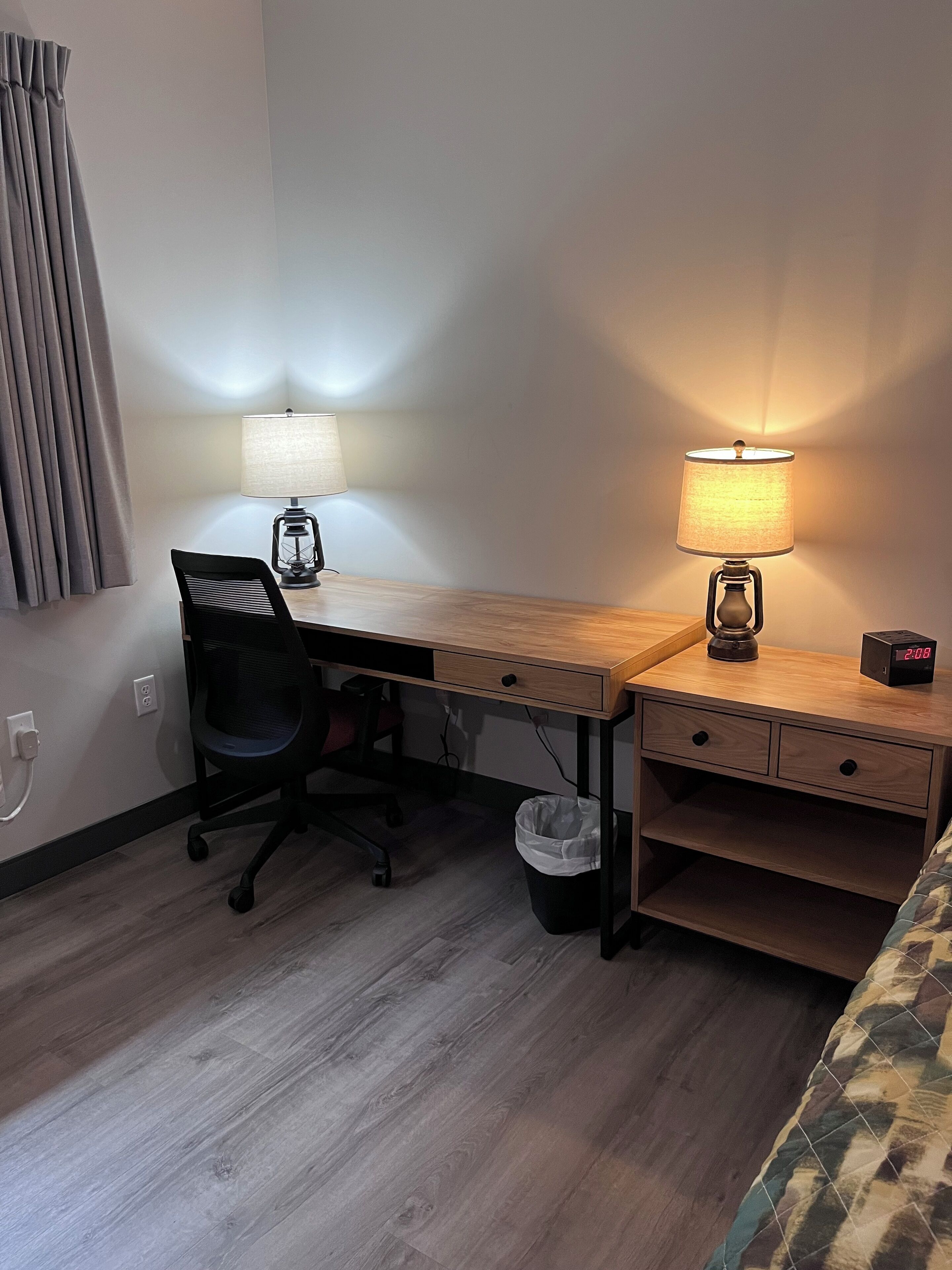 A hotel room with a wooden desk, two lamps, a chair, a bed, and a trash bin.
