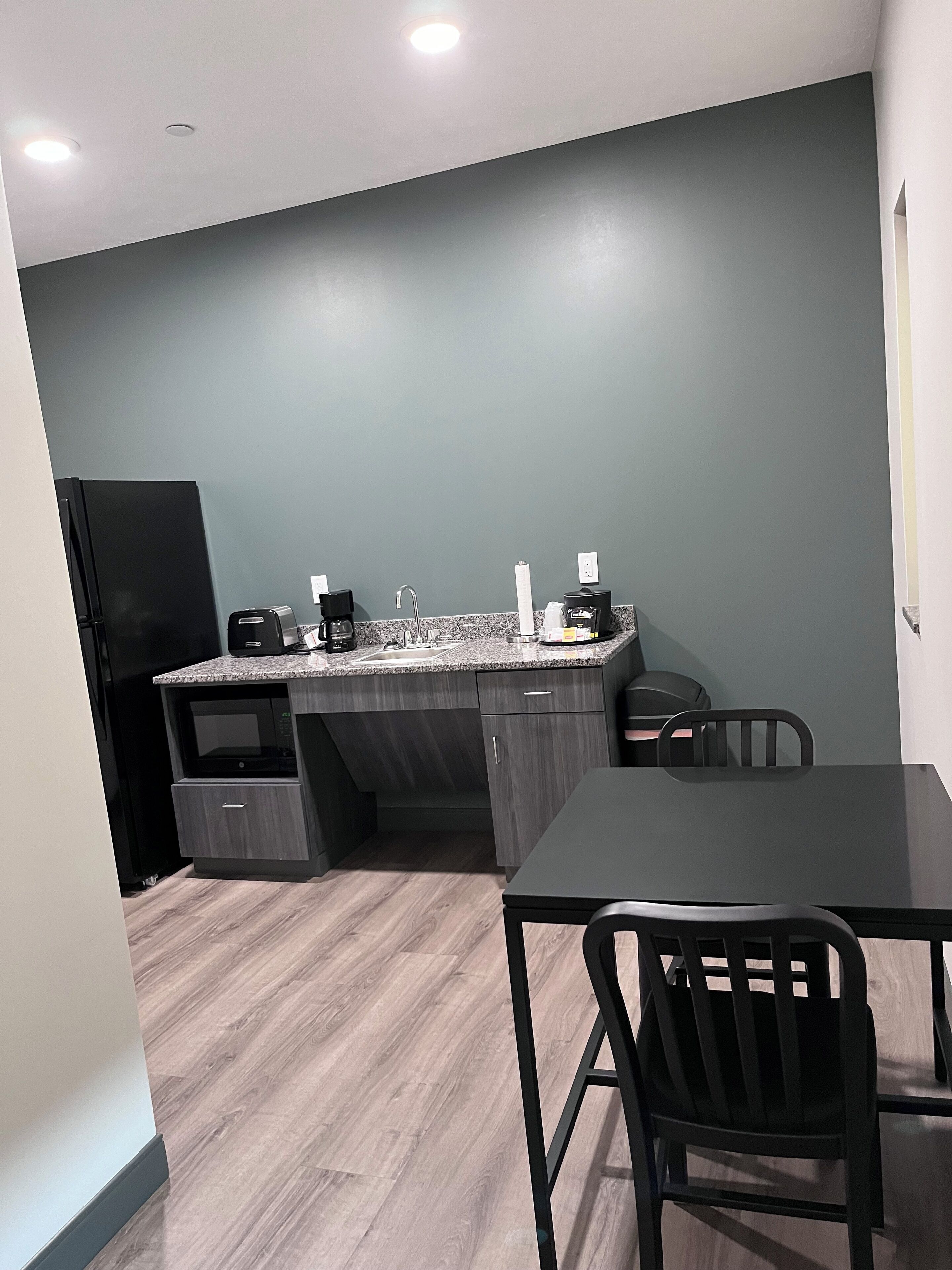A compact kitchen with a dark gray backsplash, a black refrigerator, a microwave, a coffee maker, and a sink. There is a dining table with two chairs.