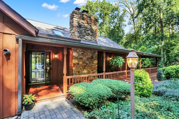 A wooden house with a stone chimney, a porch with a railing, and a garden with various plants.