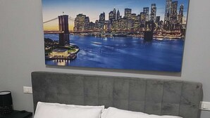 Habitación de hotel con una cama grande, un cabecero tapizado en gris y una pintura de un paisaje urbano en la pared.