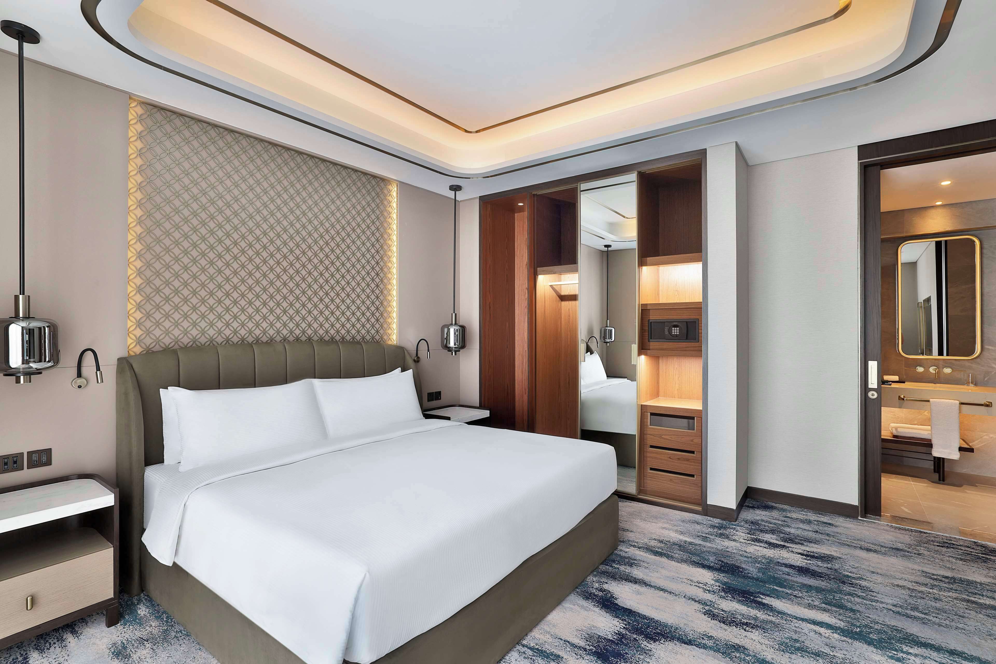 Una habitación de hotel moderna con una cama grande, mesitas de noche, un armario y un baño con espejo y lavamanos.