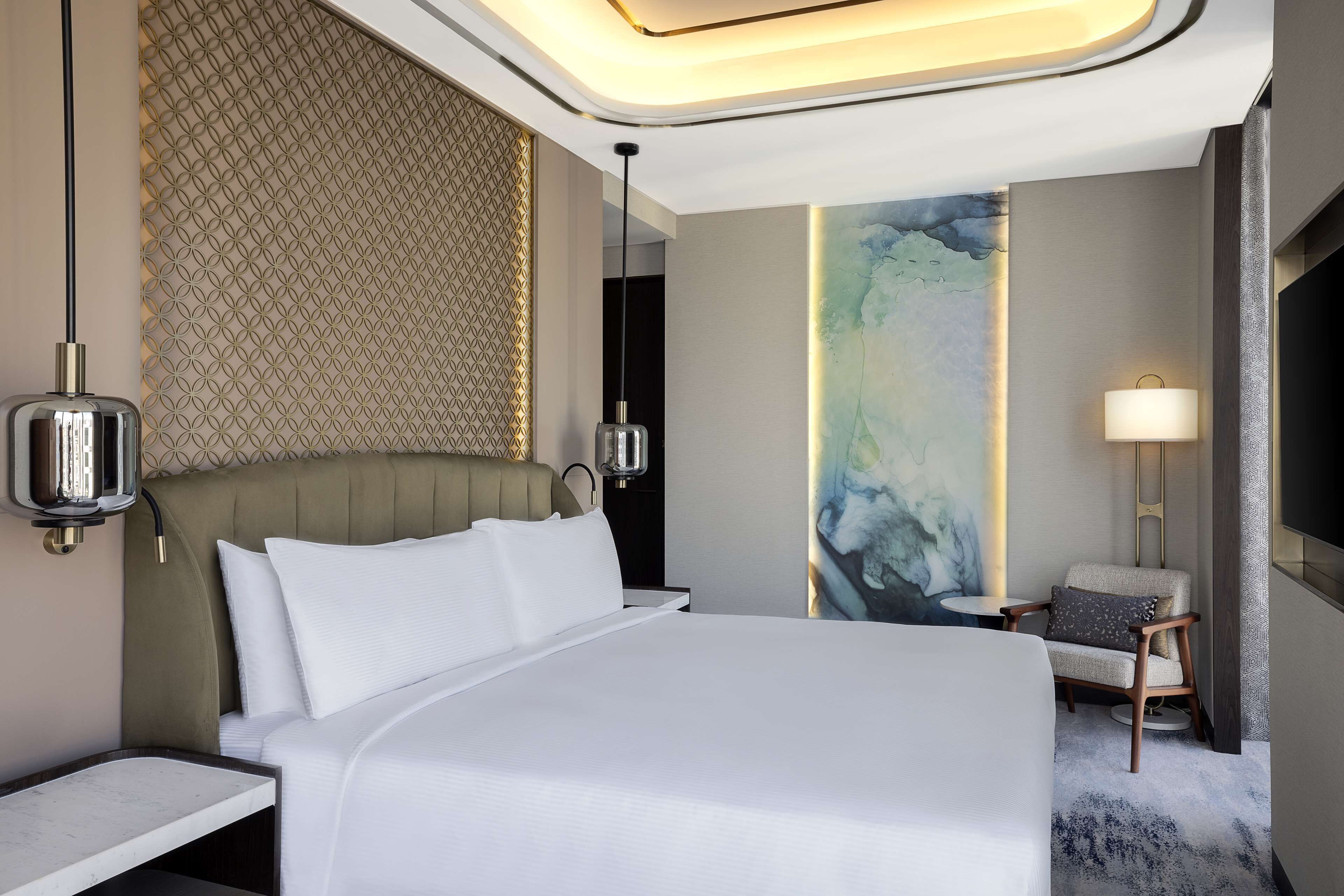 A modern hotel room with a large bed, a headboard with a geometric pattern, a side table, a chair, and a painting on the wall.