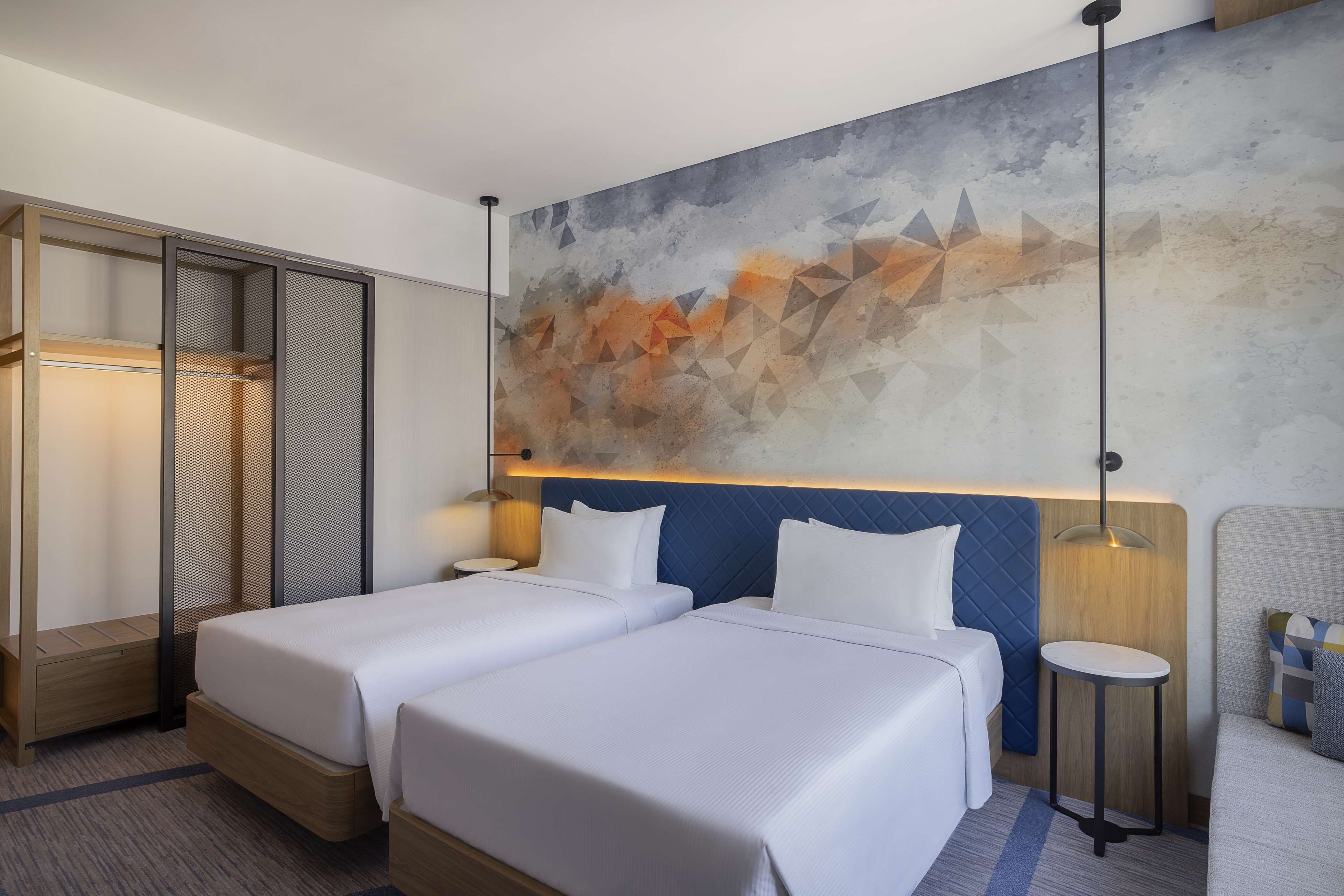 Hotellihuone, jossa on kaksi sänkyä, suuri abstrakti maalaus seinällä, puinen vaatekaappi ja pieni pyöreä pöytä, jossa on lamppu.