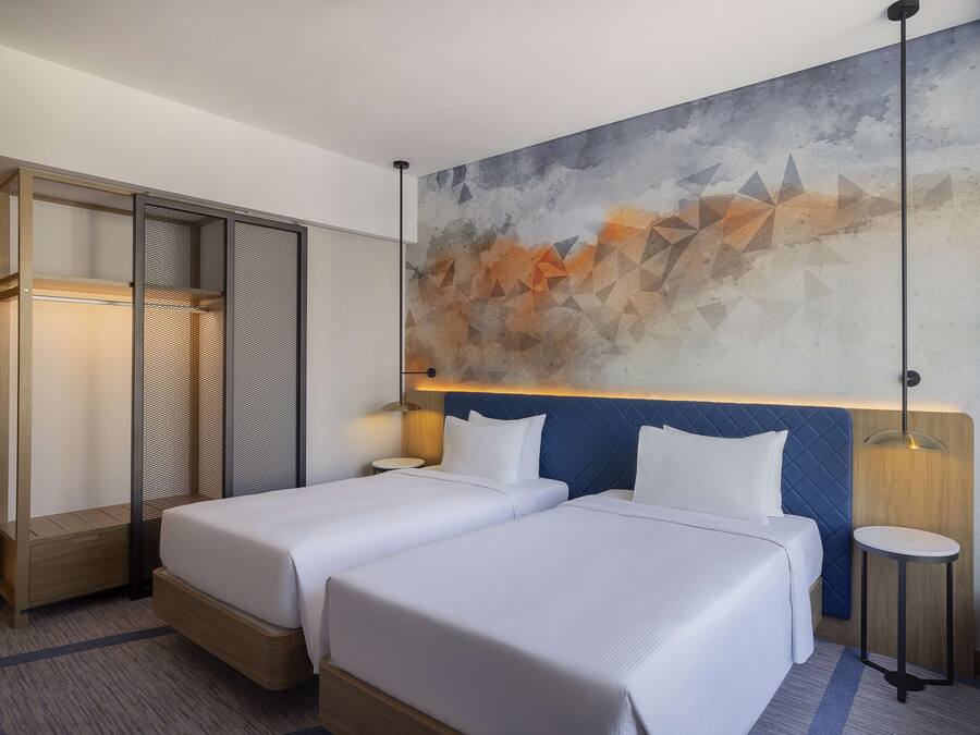 A hotel room with two beds, a large abstract painting on the wall, a wooden wardrobe, and a small round table with a lamp.