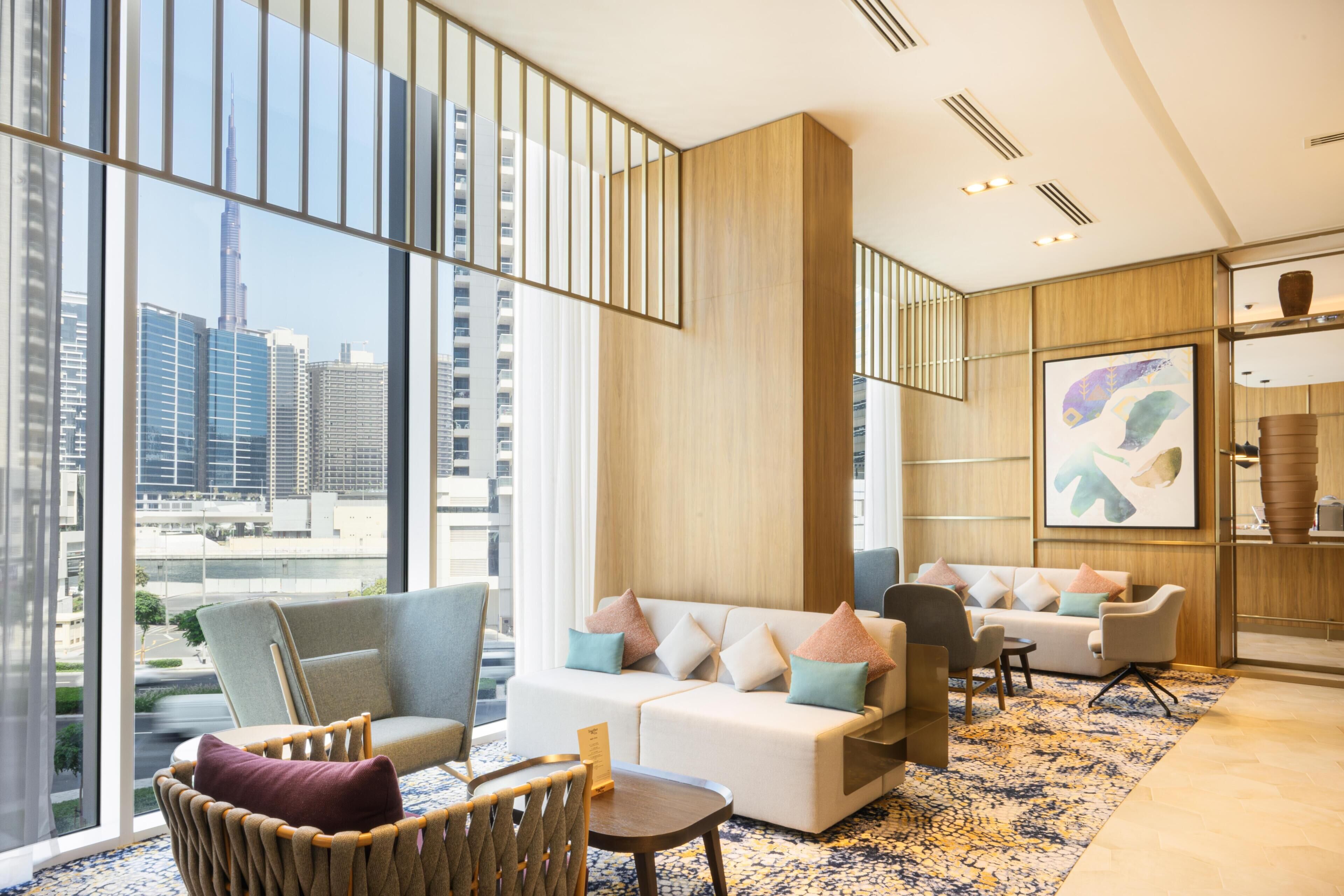 Un moderno vestíbulo de hotel con un amplio ventanal que ofrece vistas a la ciudad, un área de descanso con sofás y sillones, y un elemento decorativo en la pared.