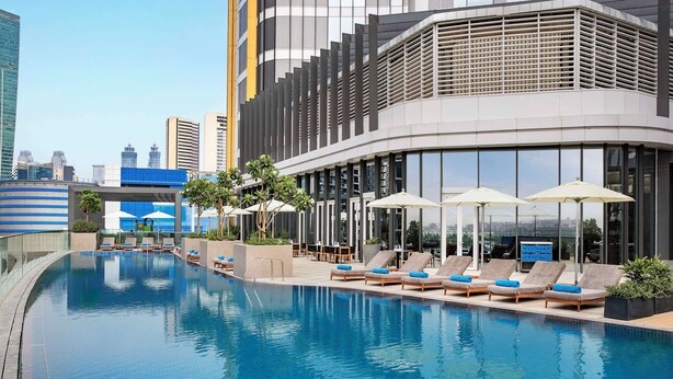 Un hotel moderno con zona de piscina que cuenta con tumbonas y sombrillas, rodeado de edificios altos.
