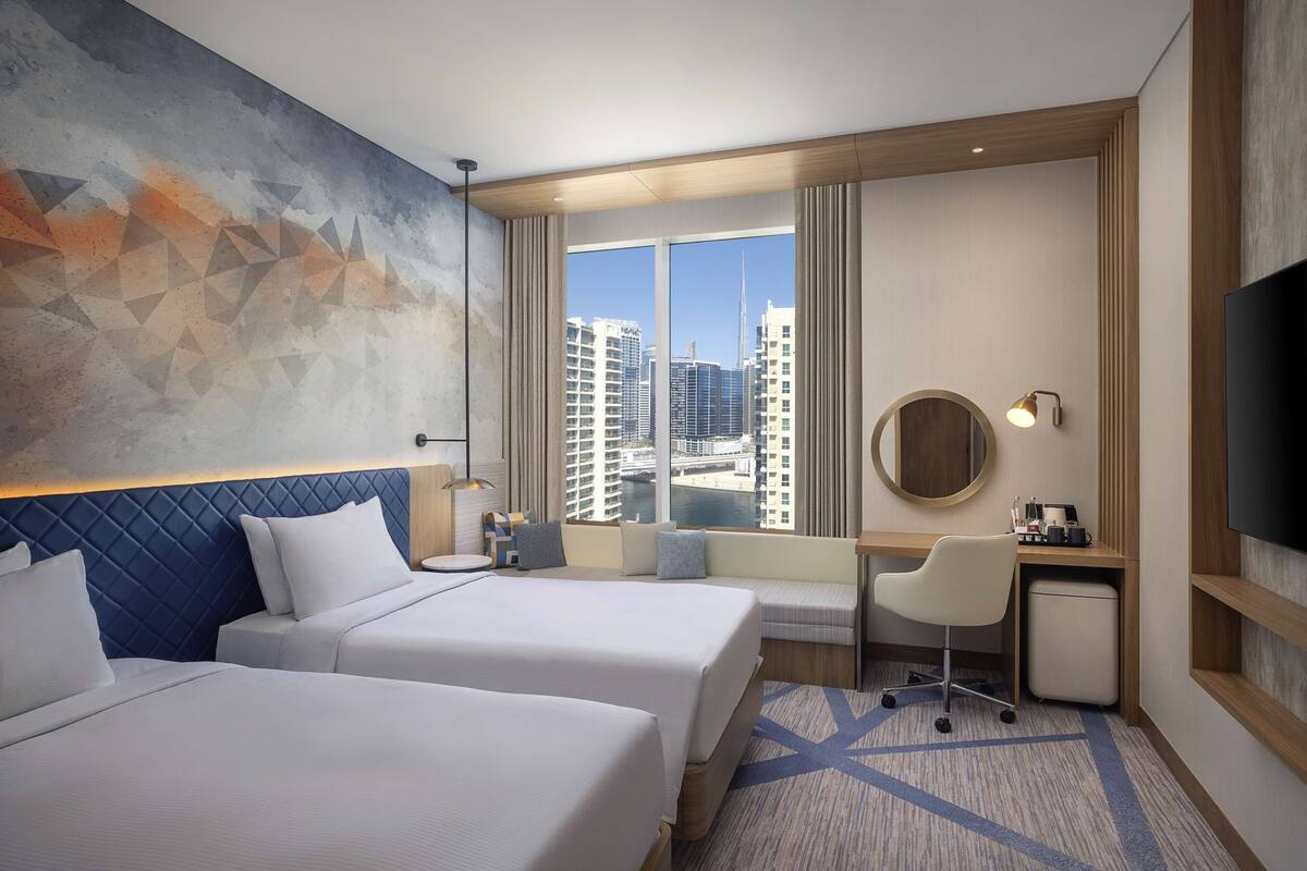 A hotel room with two beds, a desk, a chair, a TV, and a large window with a city view.