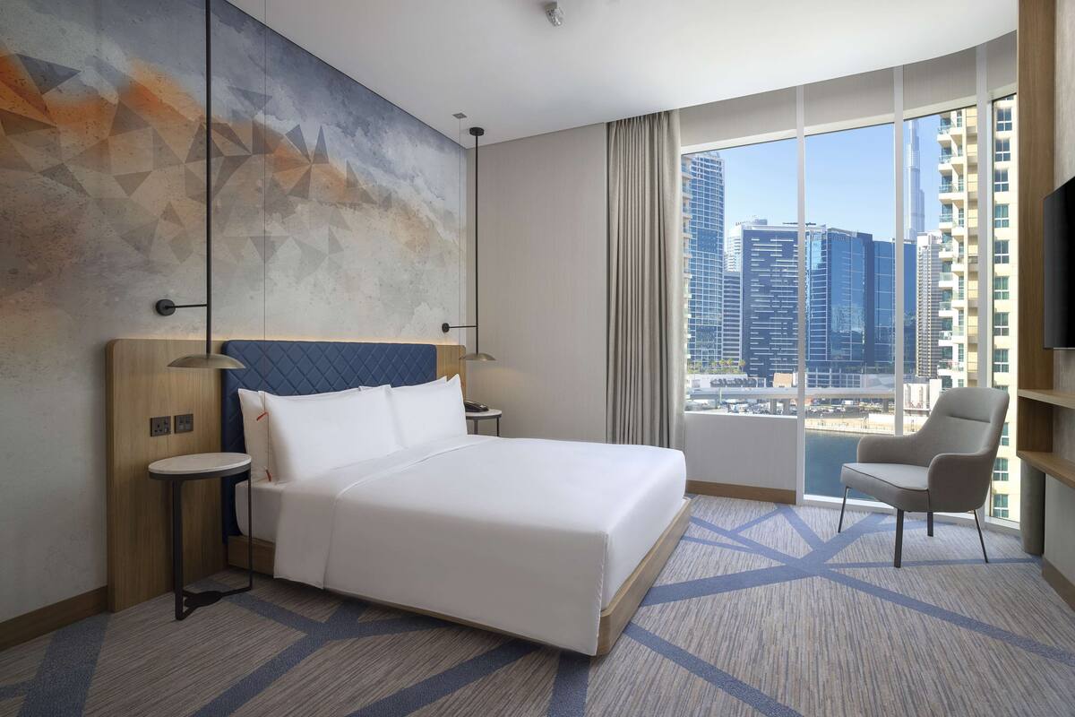 A hotel room with a large bed, a chair, a TV, and a city view.