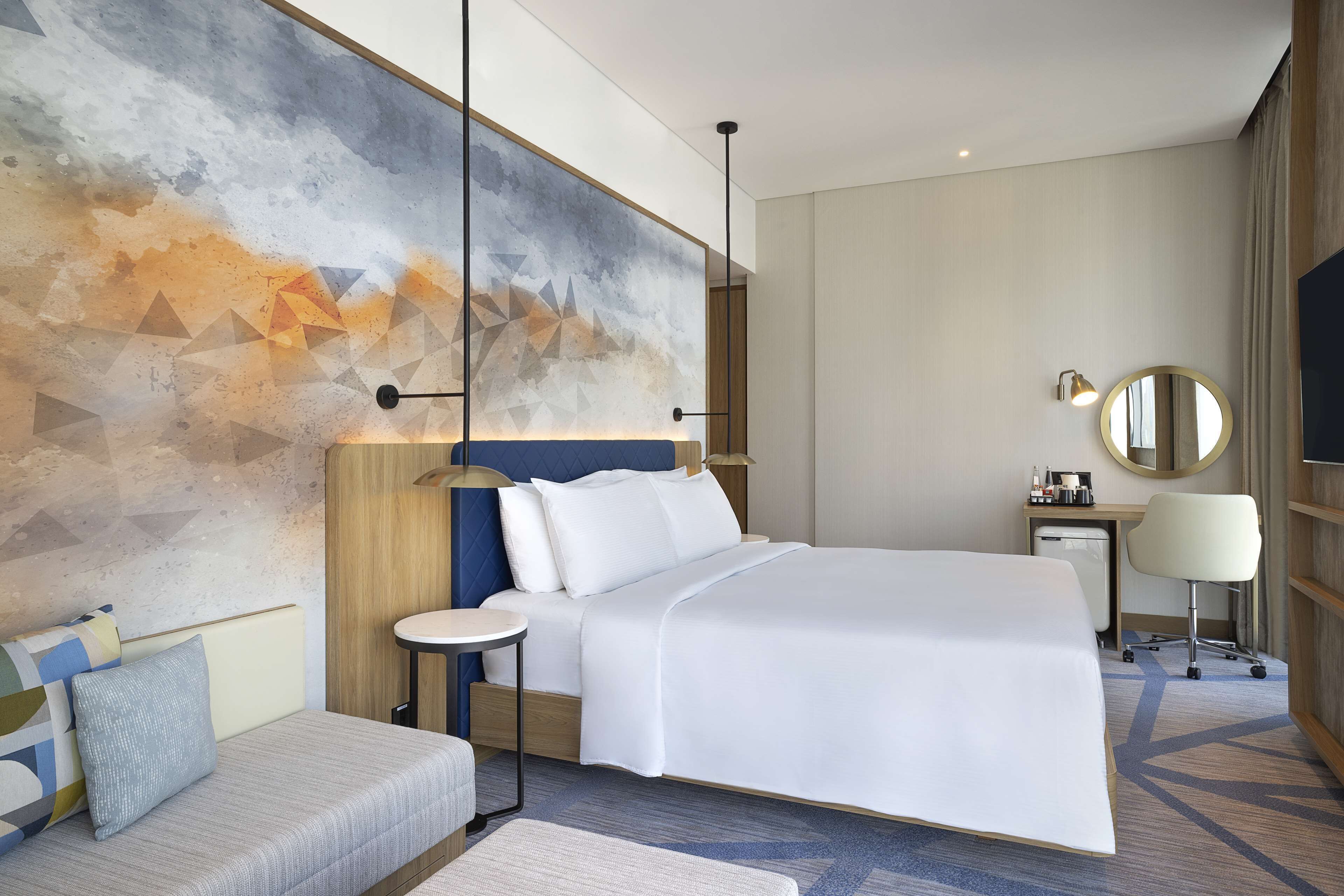 Habitació d'hotel moderna amb un llit gran, una taula de treball amb una cadira, un mirall rodó i un quadre abstracte de grans dimensions a la paret.