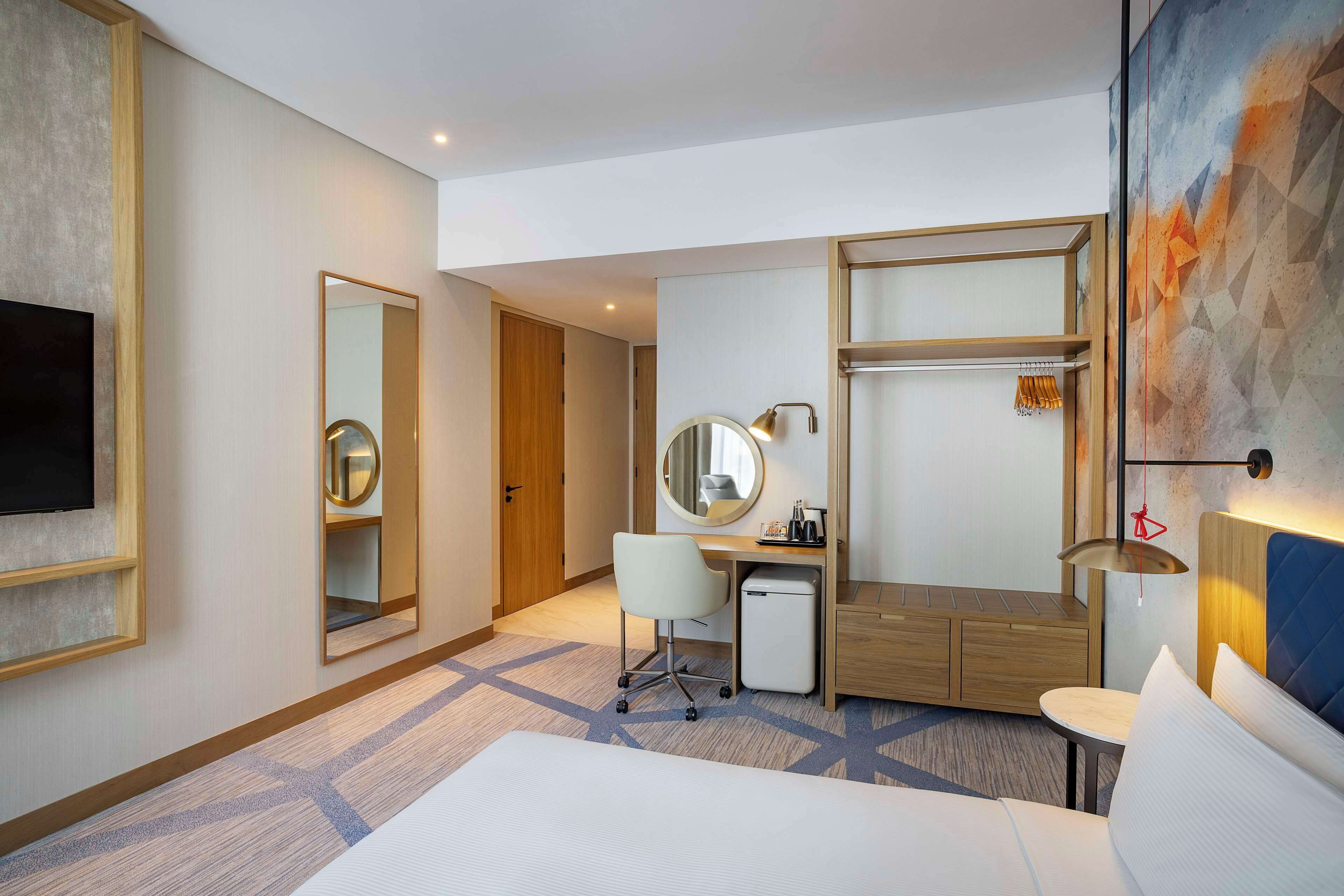 A hotel room with a bed, desk, chair, small refrigerator, and a TV.