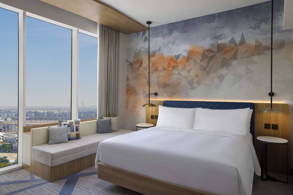 A modern hotel room with a large bed, a seating area with cushions, a city view, and a geometric wall mural.