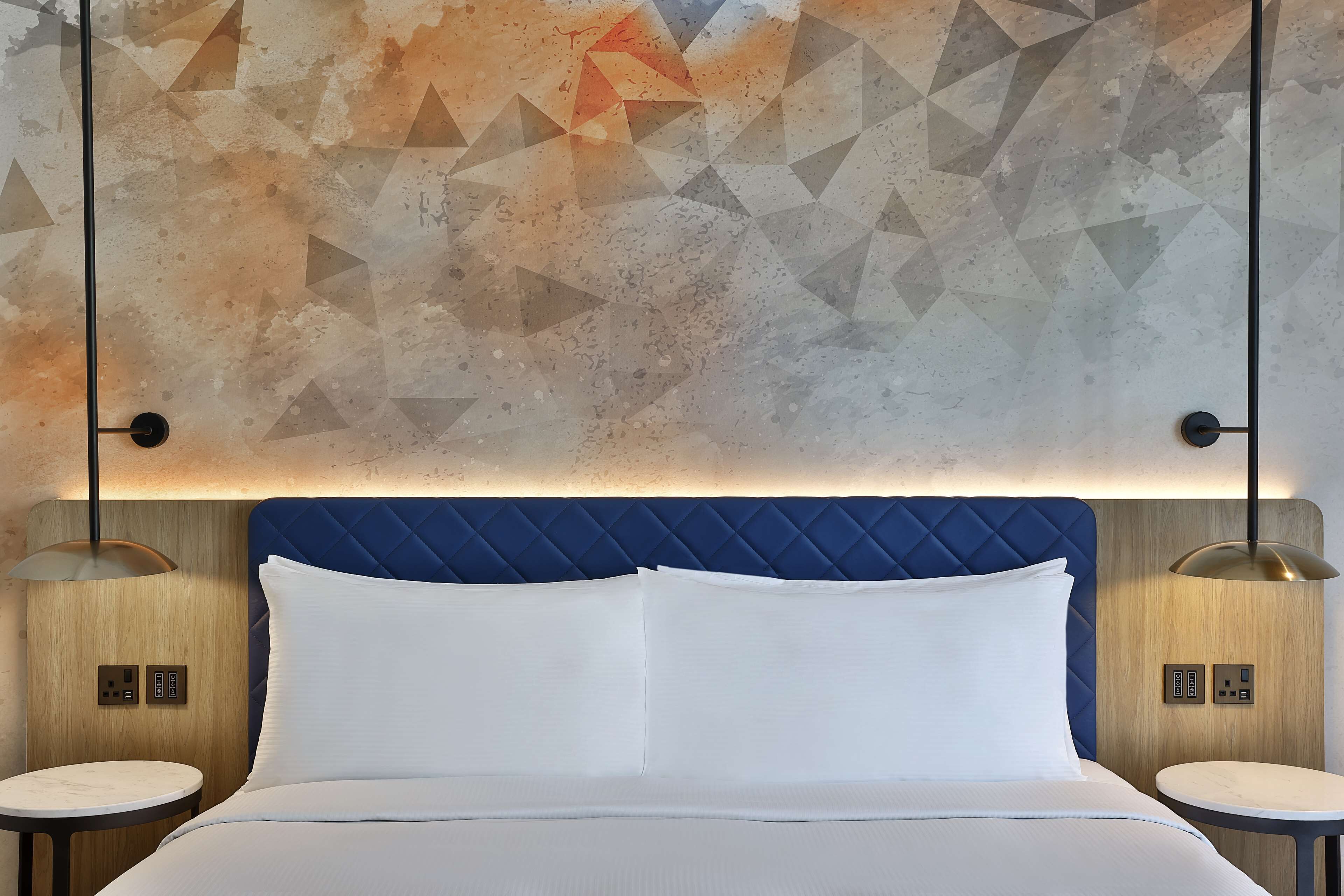 A hotel room with a bed, two bedside tables, a geometric patterned wall, and a blue headboard.