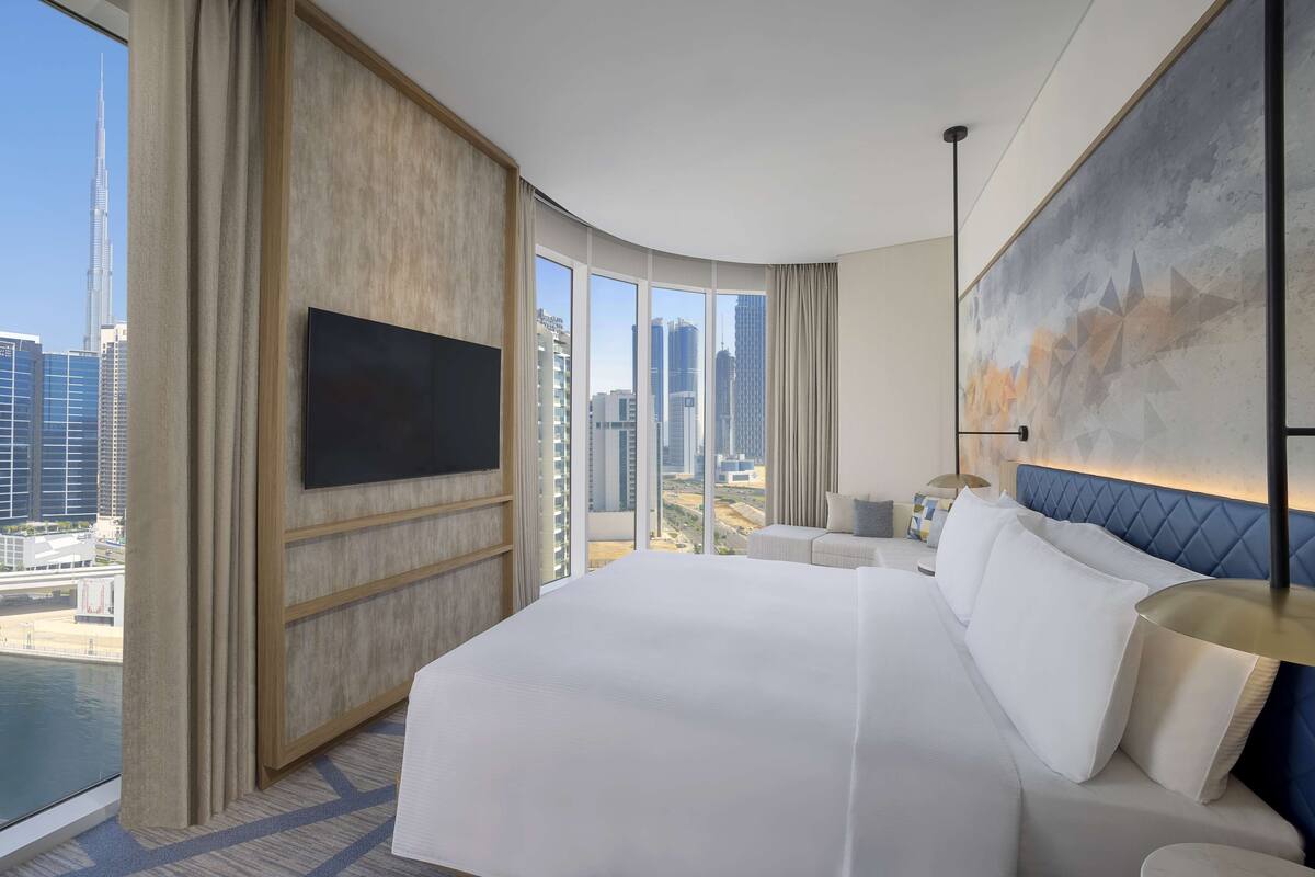 A modern hotel room with a large bed, a flat-screen TV, a city view, and a blue accent wall.