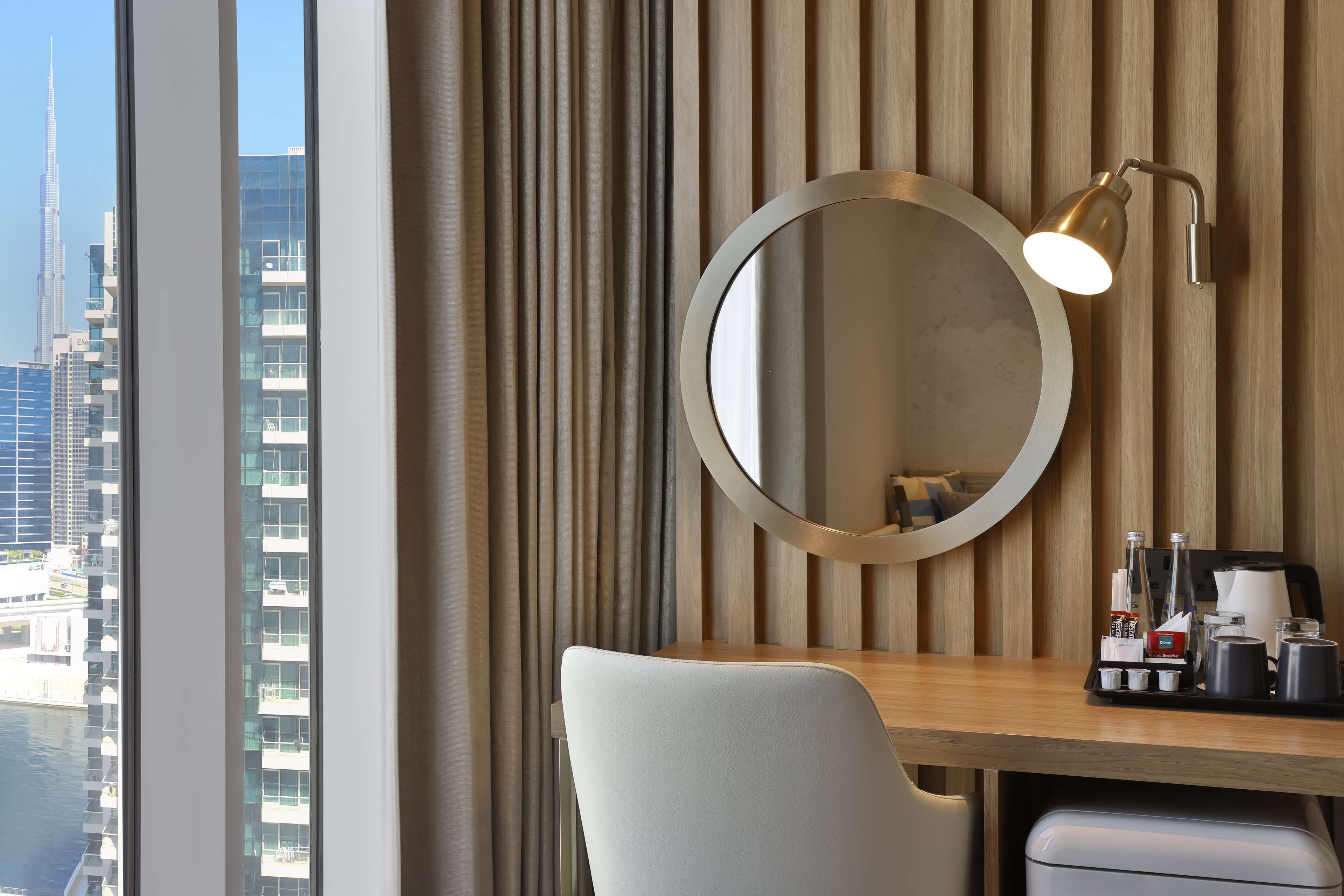 A modern hotel room with a large window, a round mirror, a desk with a coffee maker, and a trash bin.
