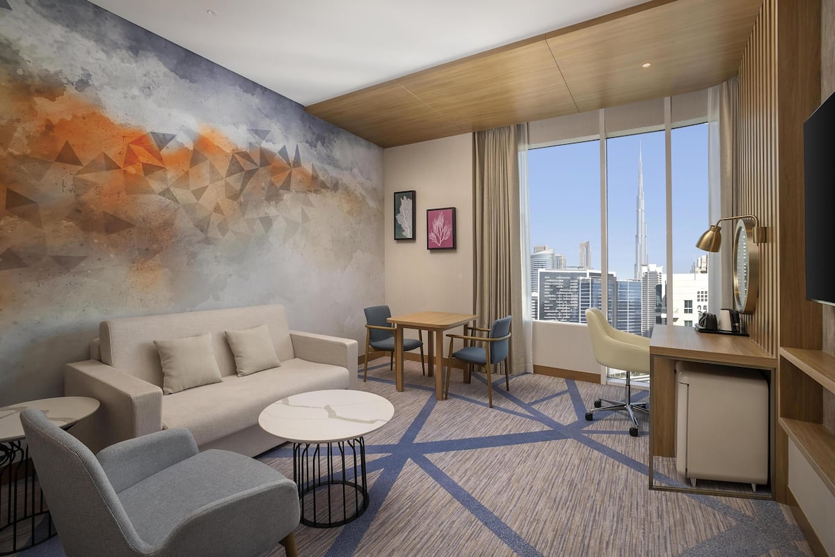 A modern hotel room with a large window offering a city view, a sofa, a small table, and a desk area.