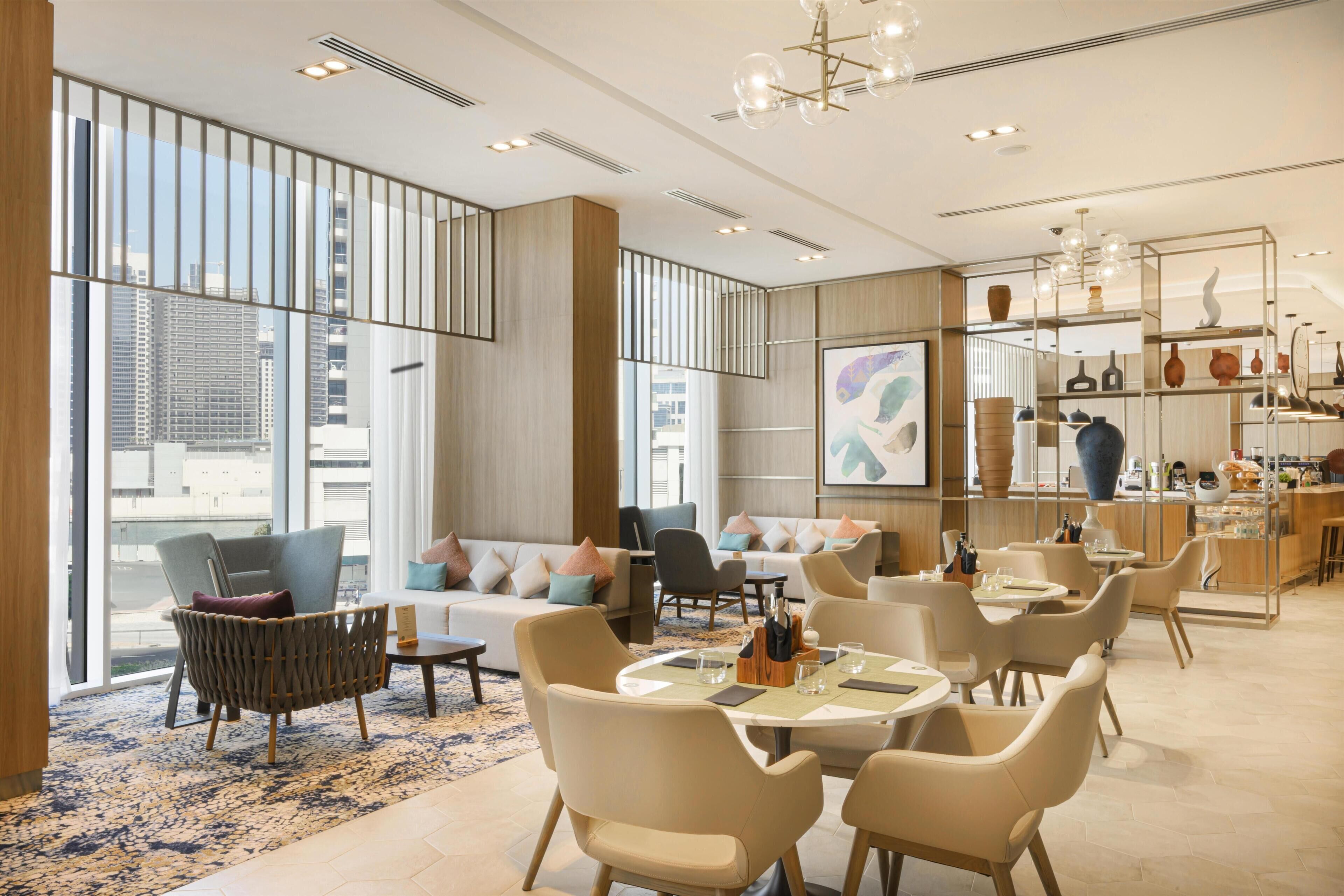 Un moderno vestíbulo de hotel con techos altos, ventanas amplias y una variedad de zonas de estar, incluyendo sofás y sillones.