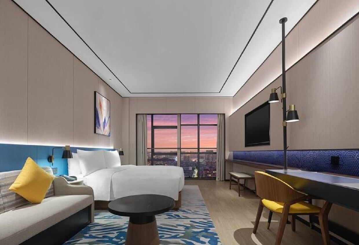 Habitación de hotel con cama, sofá, mesa de comedor, televisor y ventana grande con vista a la ciudad.