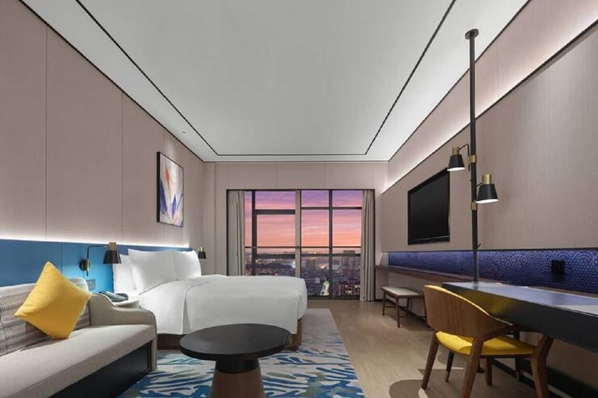Hotelkamer met een bed, een bank, een eettafel, een televisie en een groot raam met uitzicht op de stad.