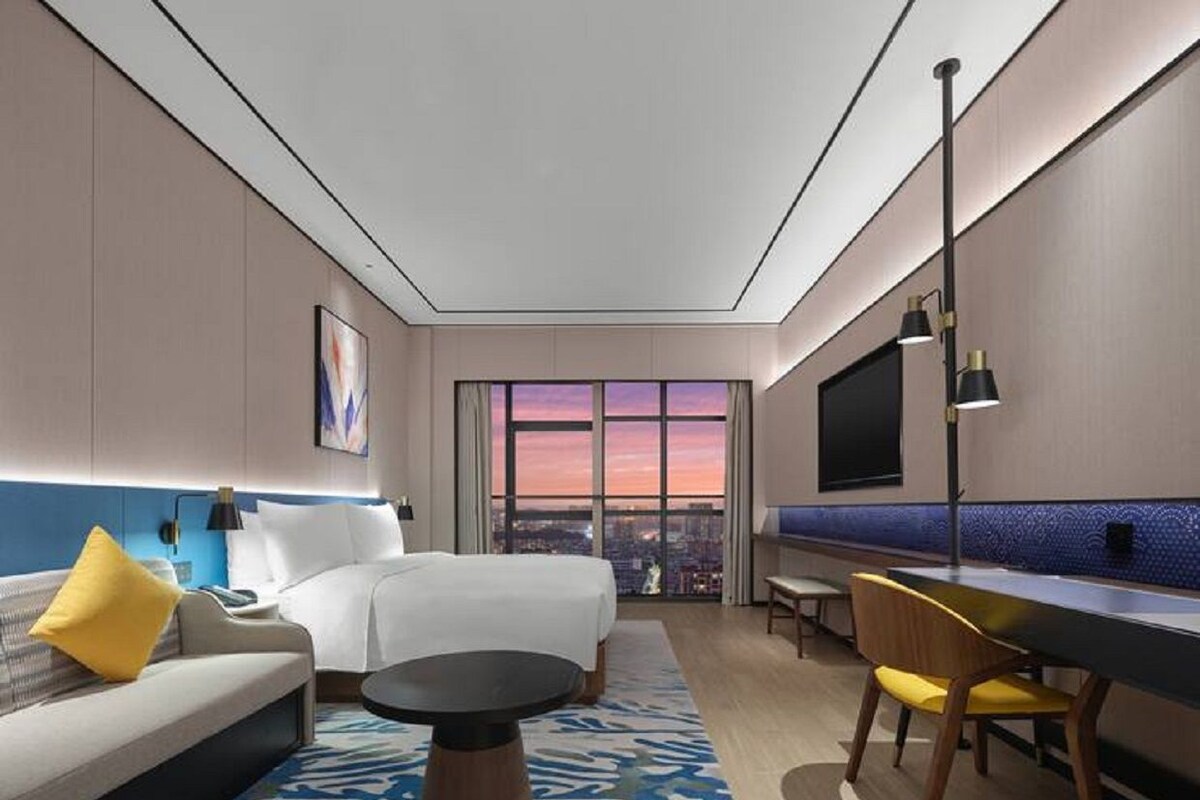 A hotel room with a bed, a sofa, a dining table, a TV, and a large window with a city view.