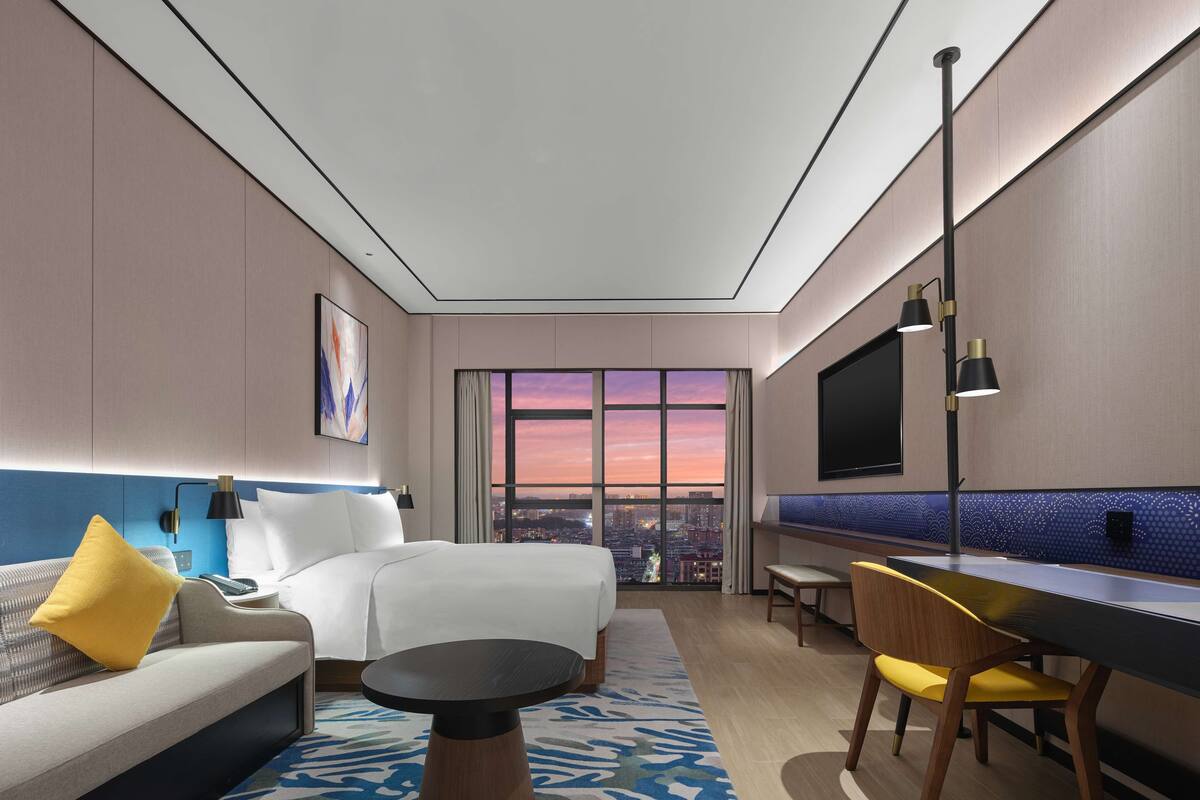 Een moderne hotelkamer met een bed, een bank, een eettafel, een stoel, een televisie en een groot raam met uitzicht op de stad.