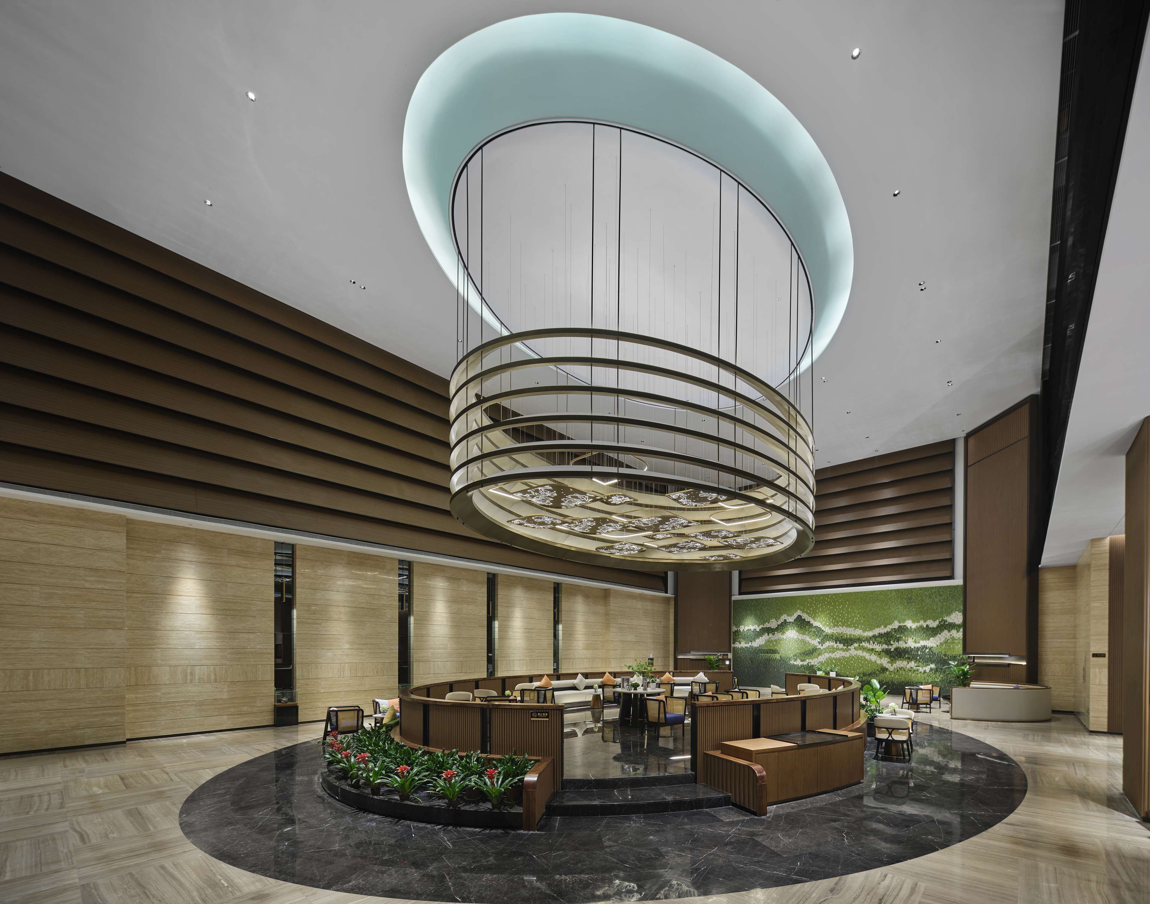 A modern lobby with a central circular reception desk, a large chandelier, and a mural on the wall.