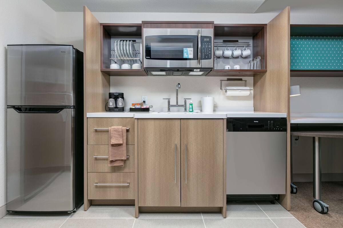 A compact kitchen with a stainless steel refrigerator, a microwave, and a dishwasher. There are wooden cabinets with a towel hanging on one of the handles.