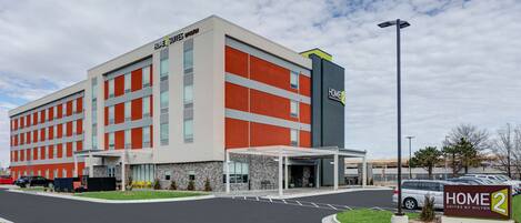 A modern hotel building with a prominent "Home 2 Suites by Hilton" sign, a covered entrance, and a parking lot.