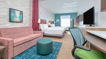 Een hotelkamer met een bed, een bureau, een televisie, een rode bank, een blauwgroene voetenbank en een kamerbreed tapijt.