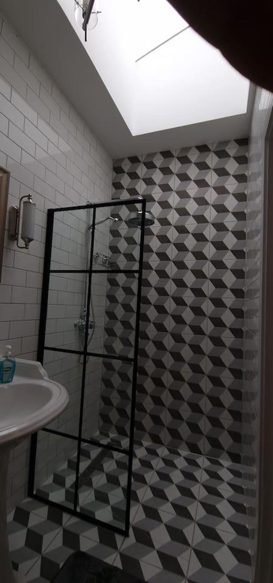 Une salle de bain avec un mur de douche orné d’un motif géométrique, une douche à parois de verre et un lavabo blanc doté d’un distributeur de savon.