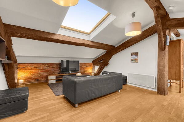 A loft-style room with exposed wooden beams, a skylight, a flat-screen TV, a grey sofa, and a brick accent wall.