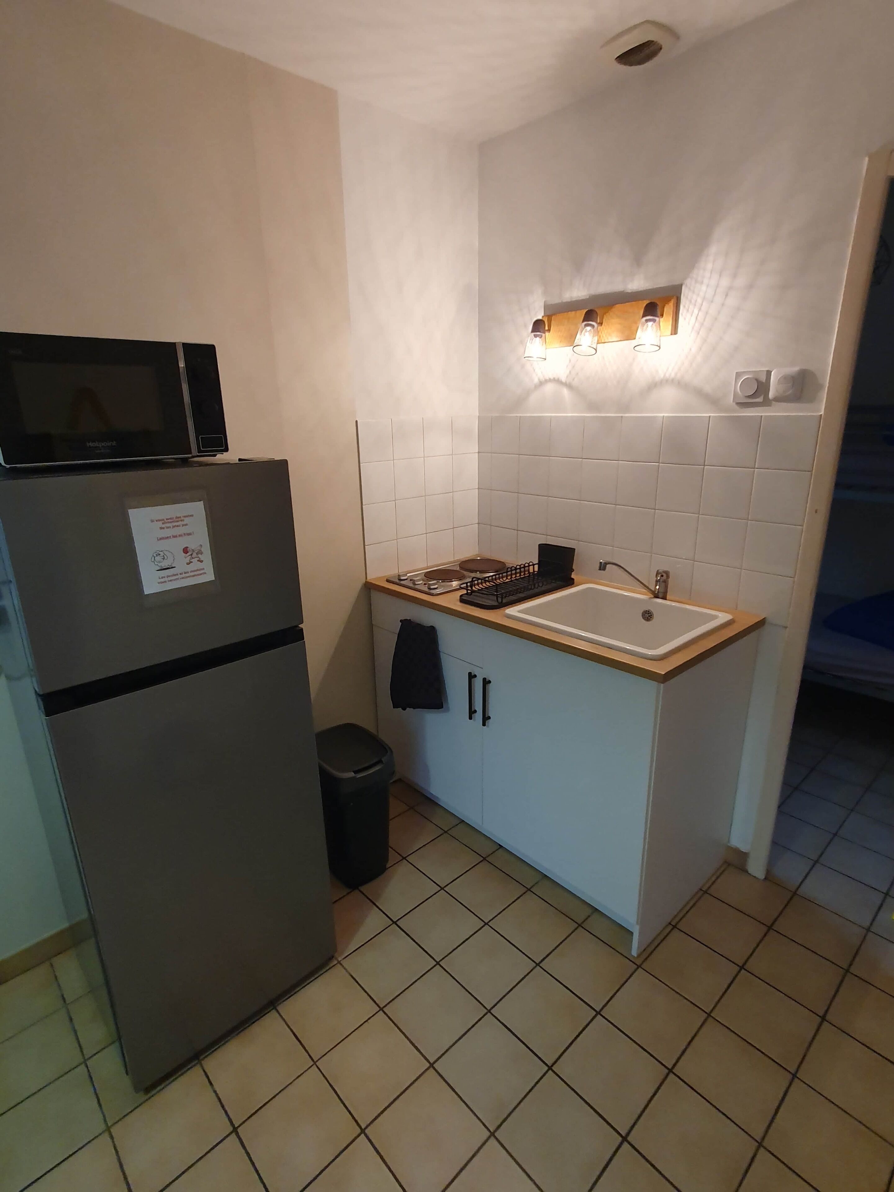 A compact kitchen with a refrigerator, stove, sink, and a wall-mounted light fixture.