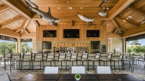 Ruime binnenruimte met een lange tafel en stoelen, houten plafond met plafondventilatoren, en een bar in de achtergrond.