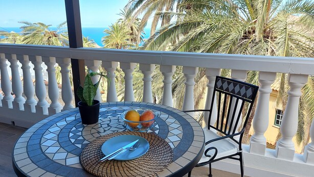 A round table with a potted plant, a bowl of fruit, and cutlery on a balcony with a white balustrade, overlooking palm trees and the sea.