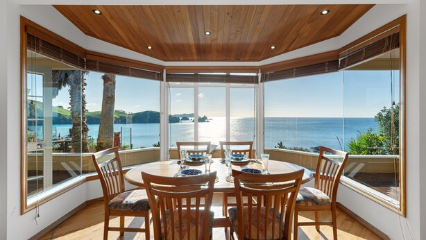 A dining area with a view of the sea, wooden ceiling, and large windows. - Seascape Point - Mahinepua Bay Clifftop Retreat (Mahinepua Bay)