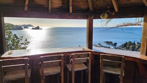 A wooden table and chairs under a thatched roof with a view of the sea and rocky coastline. - Seascape Point - Mahinepua Bay Clifftop Retreat (Mahinepua Bay)