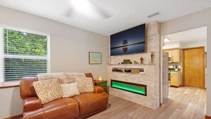A living room with a brown leather sofa, a flat-screen TV mounted on a stone wall, and a built-in fireplace with a green light. - Tree Tops Everything is brand new (St. Augustine)
