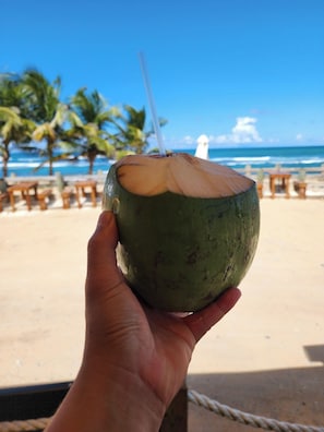 Une main tenant une noix de coco avec une paille, des palmiers et une plage en arrière-plan.