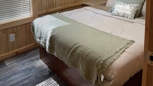A bedroom with wooden panel walls, a bed with a green blanket, a ceiling fan, and a window with blinds.