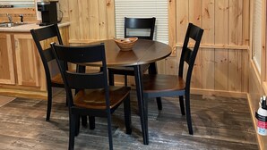 A wooden-paneled room with a dining table, four chairs, a television mounted on the wall, and a kitchen area with a sink and cabinets.