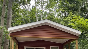 A small cabin with a wooden deck and a covered porch, surrounded by trees.