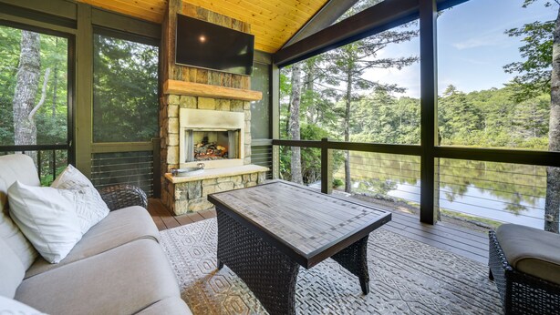 A cozy outdoor seating area with a stone fireplace, a flat-screen TV, and a view of a lake surrounded by trees. - Lakefront Gem w/ Fire Pit in Chinquapin Resort! (Glenville)