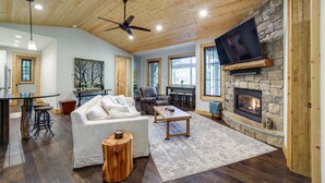 A spacious living room with a stone fireplace, wooden ceiling, and a flat-screen TV mounted above it. - Lakefront Gem w/ Fire Pit in Chinquapin Resort! (Glenville)