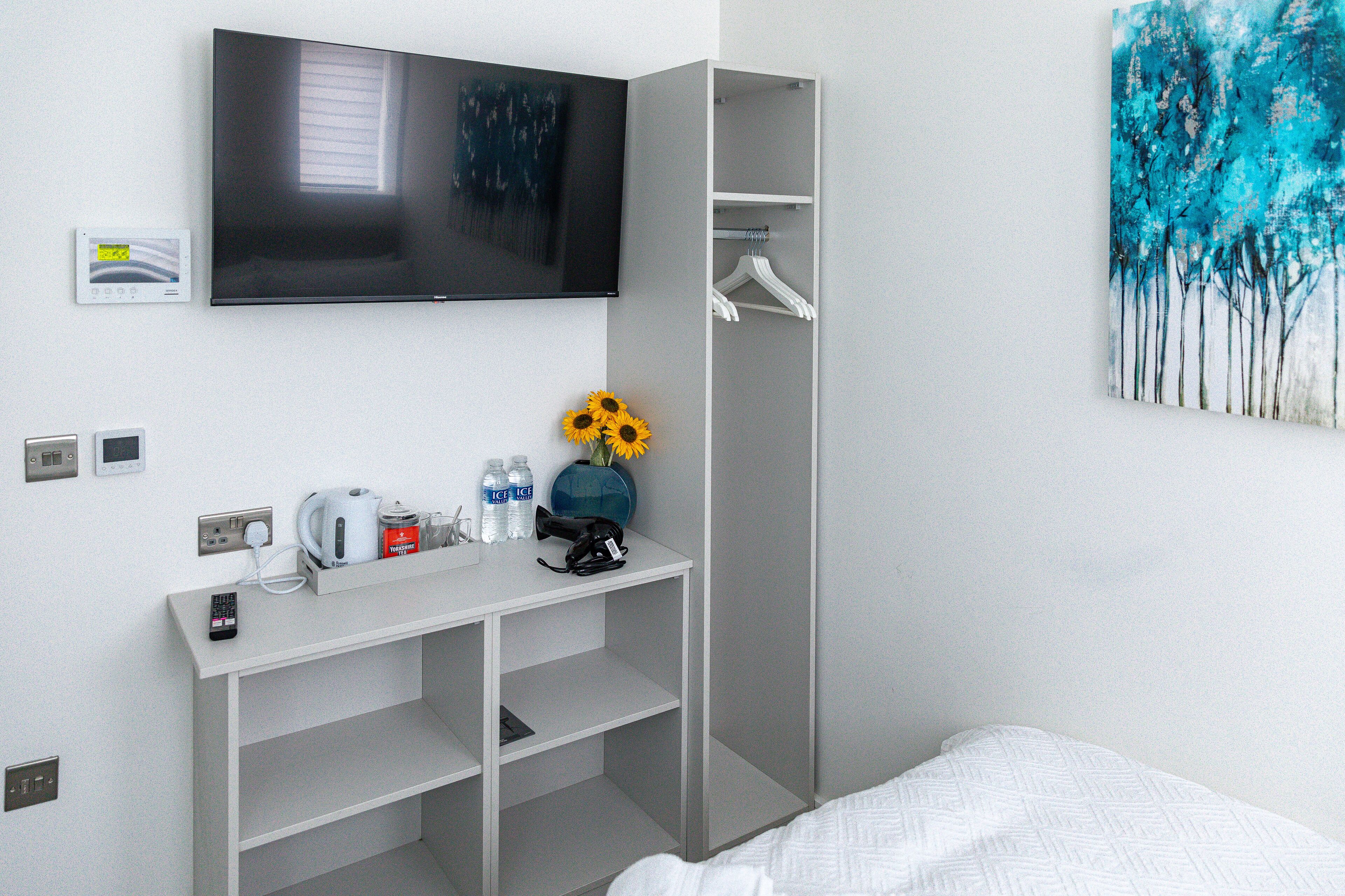 Ein Hotelzimmer mit einem Bett, einem an der Wand befestigten Fernseher, einem Schreibtisch mit Wasserkocher, Kaffeemaschine, Telefon und einer Vase mit Sonnenblumen.