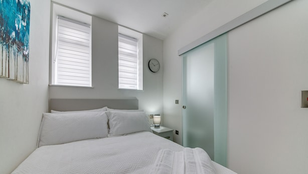 A neatly made bed with white linens, a bedside table with a lamp, a clock on the wall, and a sliding door closet.