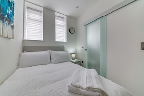 Un lit bien fait, avec du linge de lit blanc, une table de chevet avec une lampe, une horloge au mur et un placard à portes coulissantes.